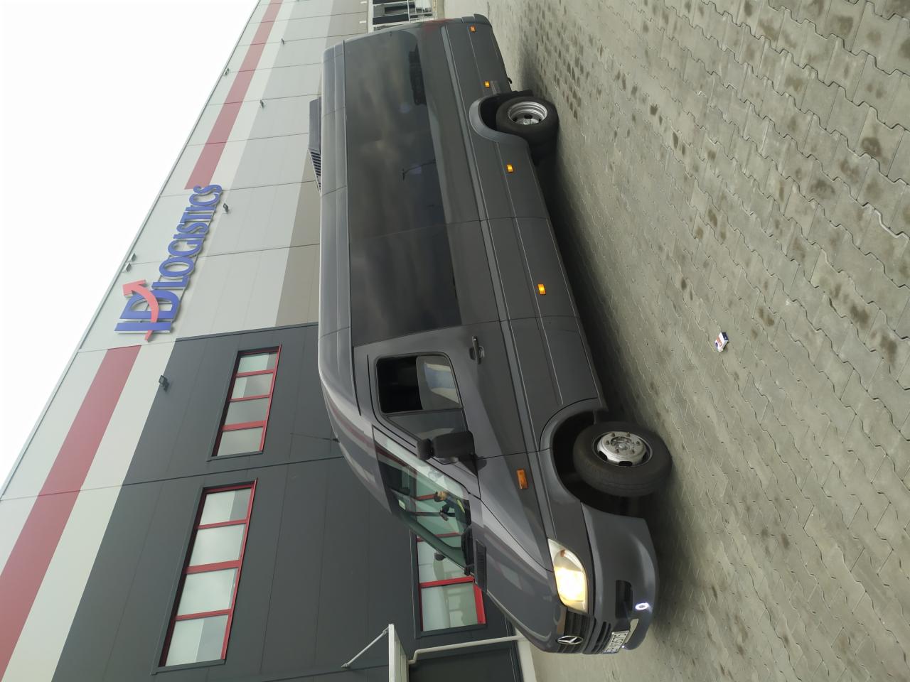 Szary bus pasażerski marki Mercedes-Benz Sprinter, zaparkowany na betonowym placu przed budynkiem z logo firmy HD Logistics.