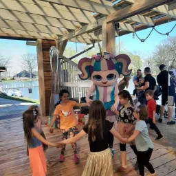 Dzieci trzymają się za ręce, tworząc koło wokół osoby w kostiumie jednorożca w Piławie Dolnej, na tle drewnianej konstrukcji z widokiem na jezioro.