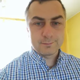 Portret mężczyzny w niebiesko-białej koszuli w kratę, z lekkim uśmiechem, na tle żółtej ściany i jasnego okna.