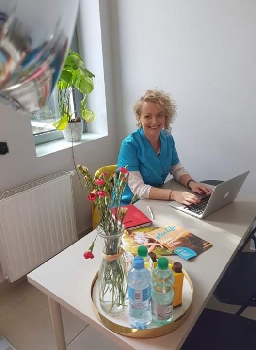 Uśmiechnięta kobieta w turkusowym uniformie siedzi przy biurku z laptopem, obok wazon z czerwonymi goździkami, butelki wody i magazyn o zdrowym stylu życia.