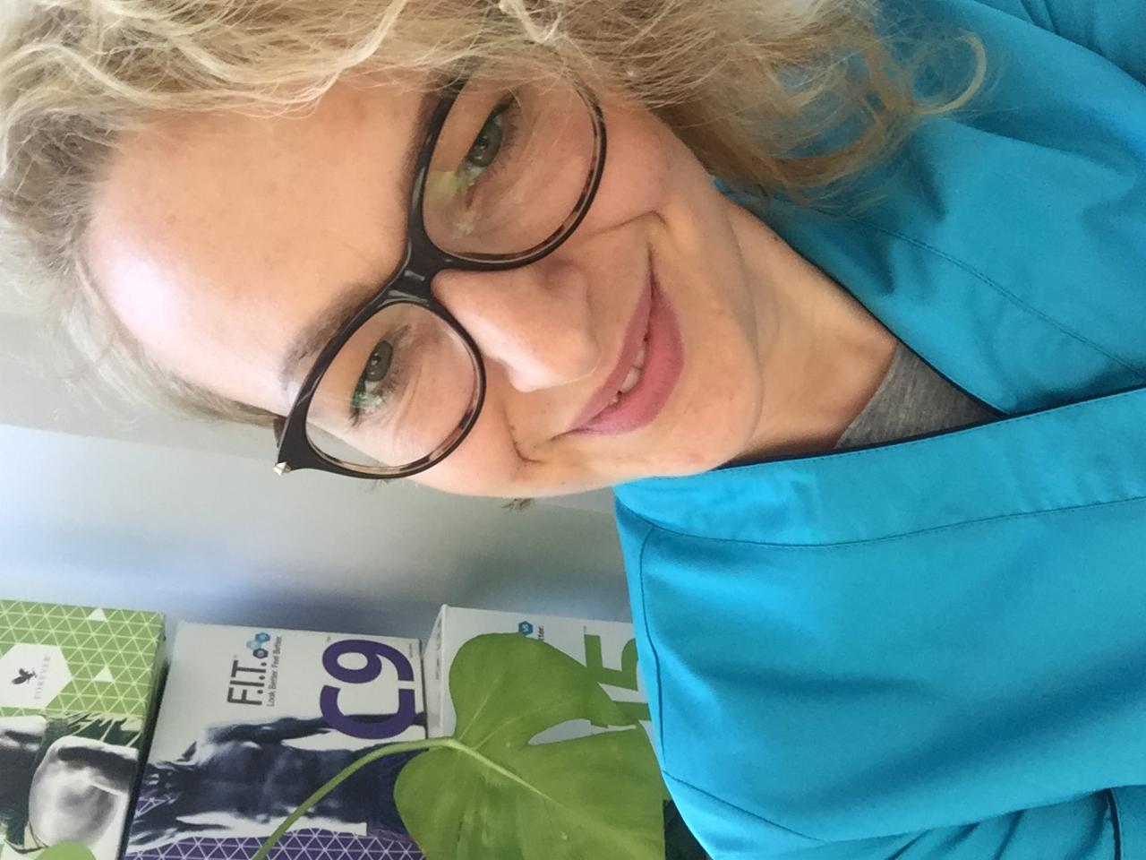 Portret uśmiechniętej kobiety w okularach i niebieskim uniformie, z widocznymi produktami F.I.T. C9 i liściem monstery w tle.