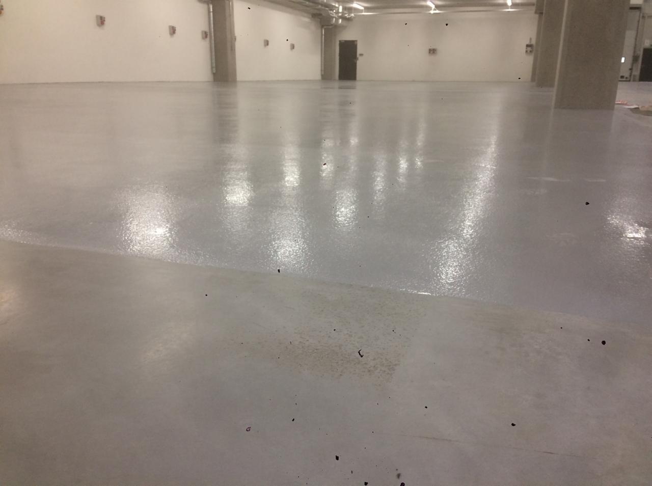 Świeżo wylana, jasnoszara posadzka w dużej hali z widocznymi kolumnami i instalacjami pod sufitem.