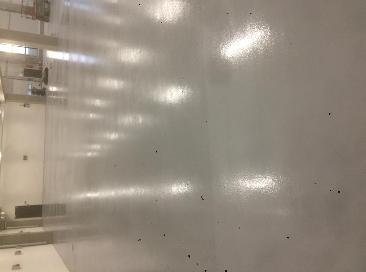 Błyszcząca, szara posadzka w hali przemysłowej, widoczne refleksy świetlne i drobne zabrudzenia.