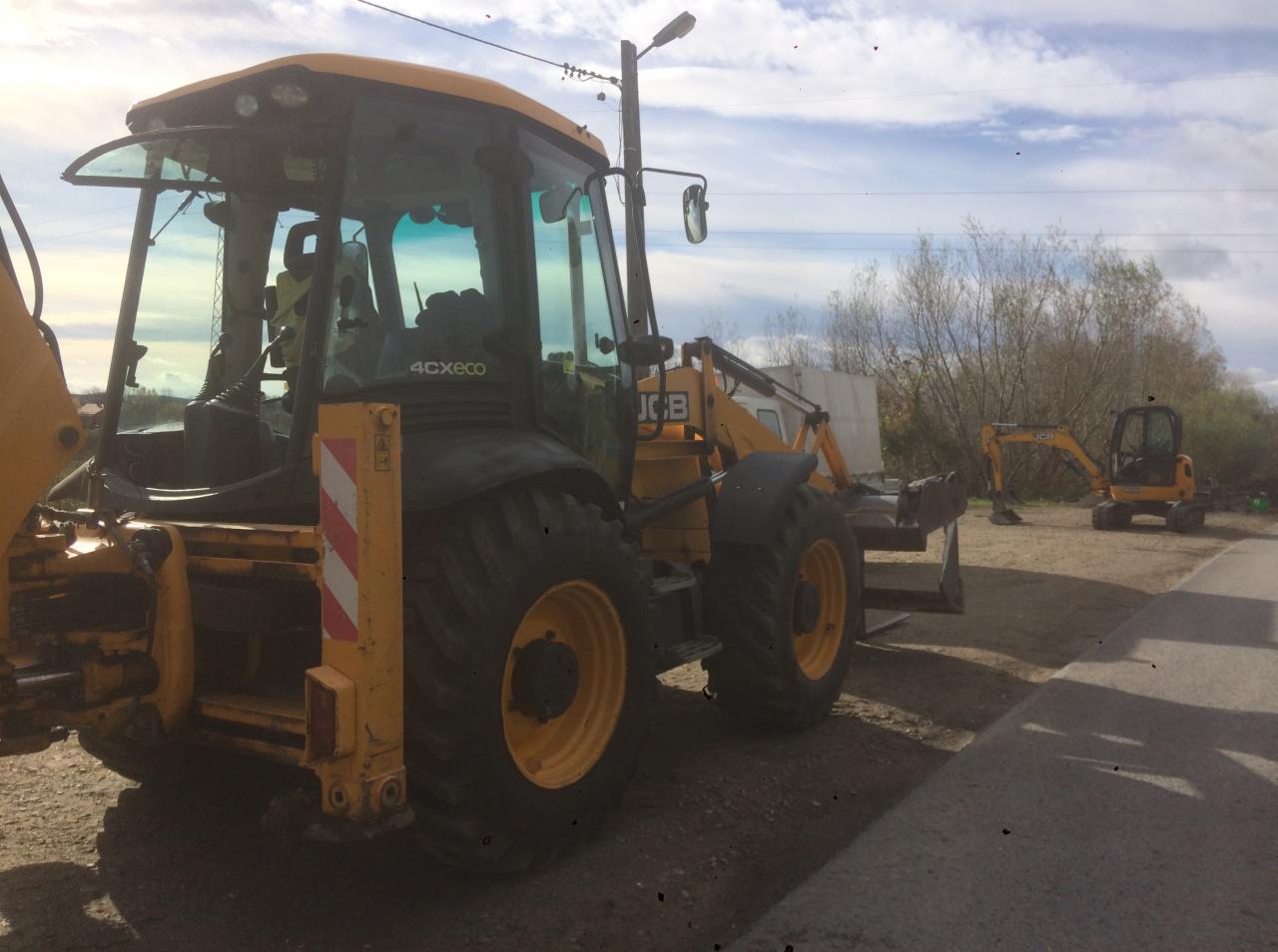 Żółto-czarna koparko-ładowarka JCB 4CXeco z widocznymi oznaczeniami bezpieczeństwa, stojąca na placu budowy z drugą, mniejszą koparką w tle, pod błękitnym niebem z chmurami.