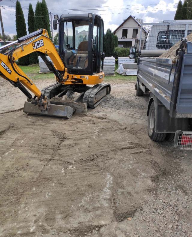 Żółta mini koparka JCB na placu budowy, obok zaparkowana przyczepa z piaskiem, w tle dom jednorodzinny.