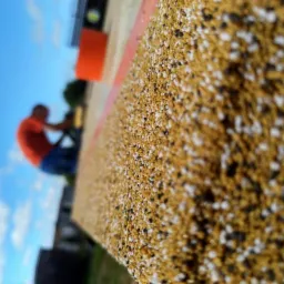 Zbliżenie na powierzchnię z żywicy epoksydowej z drobnymi kamyczkami w odcieniach beżu, bieli i czerni, z rozmytą sylwetką mężczyzny pracującego w tle na tle nieba i zieleni.