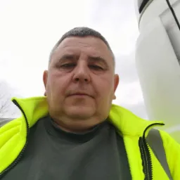 Kierowca w neonowej kurtce na tle białej kabiny ciężarówki. Widok z dołu, pochmurne niebo w tle. Mężczyzna o krótkich włosach patrzy w obiektyw.