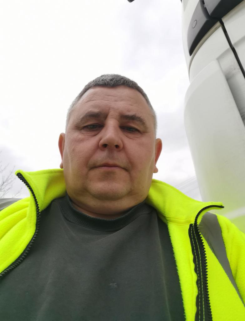 Kierowca w neonowej kurtce na tle białej kabiny ciężarówki. Widok z dołu, pochmurne niebo w tle. Mężczyzna o krótkich włosach patrzy w obiektyw.