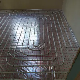 Białe rury ogrzewania podłogowego ułożone w pętle na folii z czerwoną siatką w jasnym pomieszczeniu, widok z góry pod kątem.