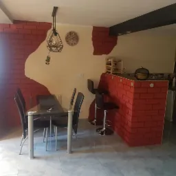 Jadalnia z elementami dekoracyjnymi imitującymi czerwoną cegłę na ścianie i barze, szklany stół z czarnymi krzesłami, nowoczesna lampa wisząca, jasne ściany i szara podłoga.