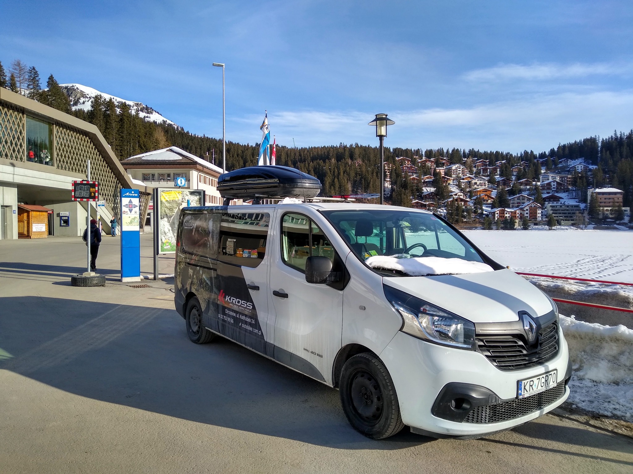 Biały van z czarnym bagażnikiem dachowym na tle zimowego krajobrazu górskiego w Arosa, Szwajcaria, z widoczną reklamą firmy Kross na boku pojazdu.