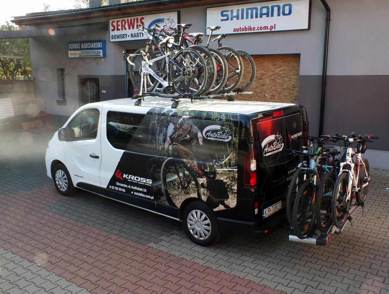 Biało-czarny bus z rowerami na dachu i z tyłu, zaparkowany w Chrzanowie przed serwisem rowerowym i narciarskim z logo Shimano i Kross na karoserii.