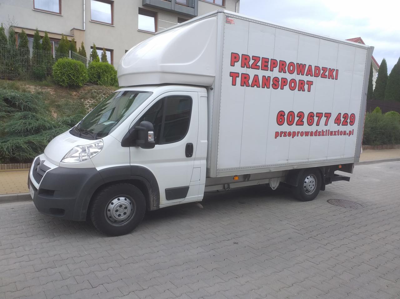Biały bus z nadrukiem 'Przeprowadzki Transport 602 677 429 przeprowadzkiluxton.pl' na tle budynku mieszkalnego i zieleni.