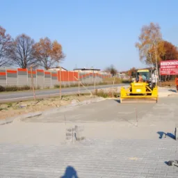 Żółty walec drogowy ugniata świeżo wylany beton na przygotowanej podbudowie pod kostkę brukową, w tle bariera akustyczna i billboard reklamowy.