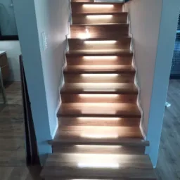 Drewniane schody z wbudowanym oświetleniem LED na każdym stopniu, widok z dołu, z fragmentem dywanika na pierwszym planie.