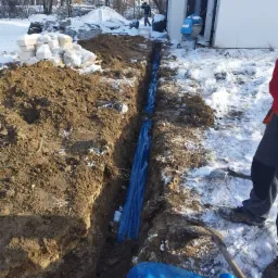 Wąski wykop w ziemi zabezpieczony niebieską folią, obok budynku w zimowej scenerii. Na pierwszym planie fragment łopaty i butów robotnika, w tle sterta bloczków.