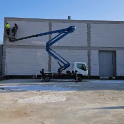 Dwóch pracowników w koszu podnośnika teleskopowego, pracujących przy szczycie murowanego budynku z betonu komórkowego w słoneczny dzień.