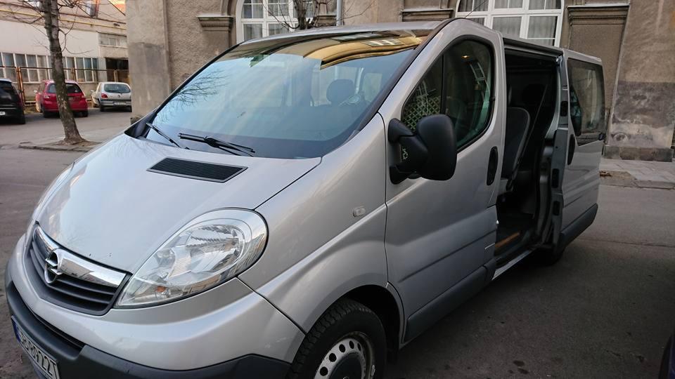 Srebrny van Opel Vivaro z otwartymi przesuwnymi drzwiami, zaparkowany na ulicy w pobliżu budynku.