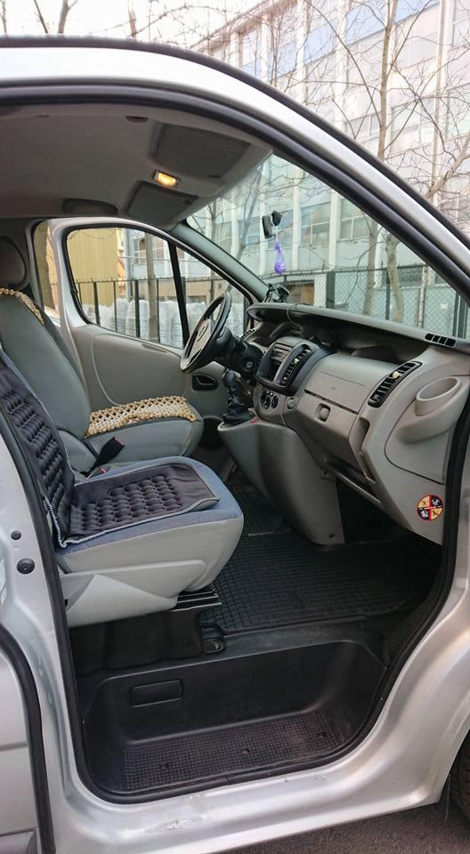 Wnętrze srebrnego busa z widocznym fotelem kierowcy z poduszką masującą, deską rozdzielczą i widokiem na budynek przez szybę.