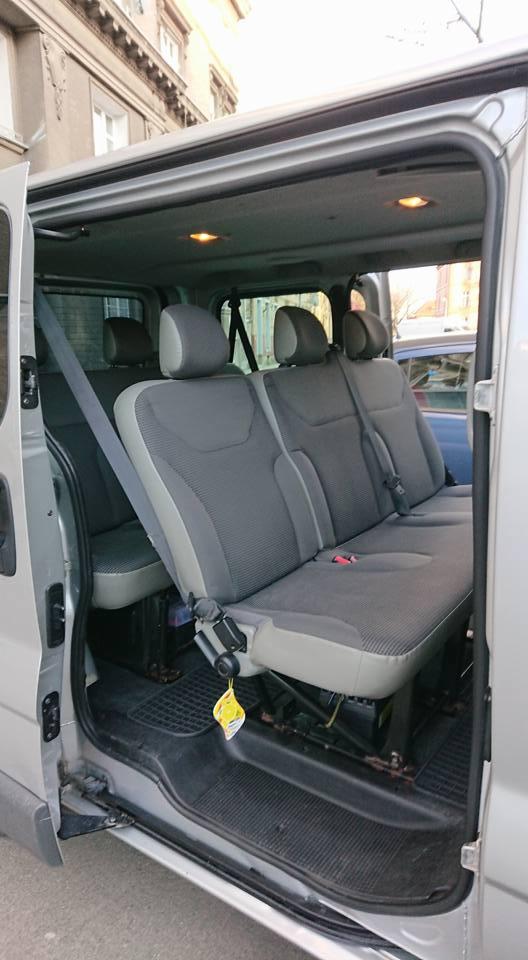 Wnętrze srebrnego busa z otwartymi drzwiami, widoczne szare fotele z pasami bezpieczeństwa i gumowa wycieraczka pod nogami.