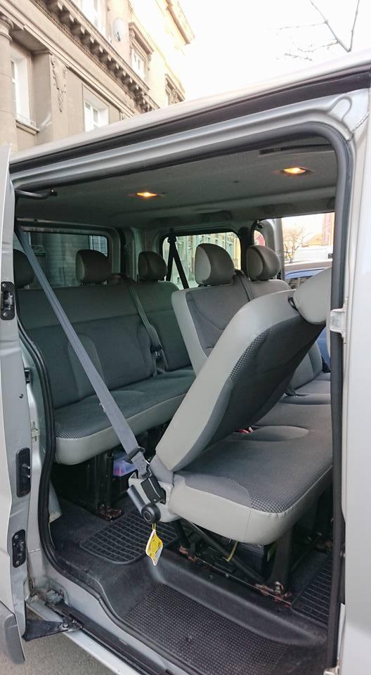 Wnętrze srebrnego busa z widocznymi fotelami pasażerskimi, jeden fotel odchylony do przodu z żółtą zawieszką, widok z otwartych drzwi bocznych na ulicę.