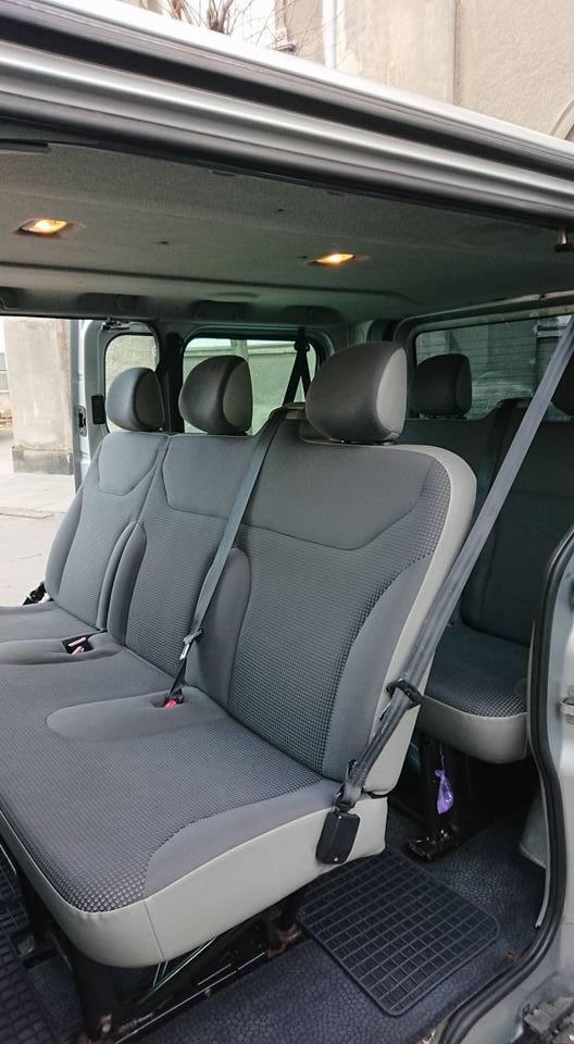 Wnętrze srebrnego busa z trzema rzędami szarych foteli, widok z boku na tylne siedzenia z pasami bezpieczeństwa, oświetlenie sufitowe.