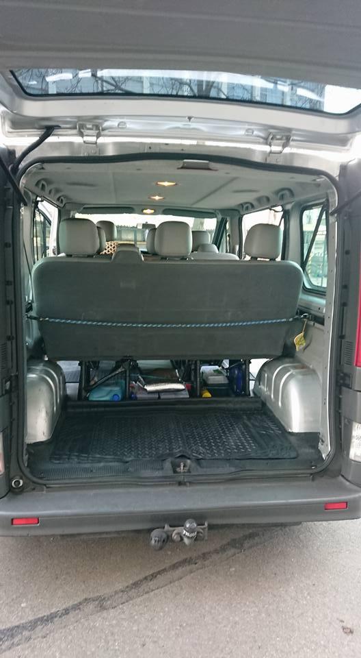 Wnętrze busa z widocznymi siedzeniami i przestrzenią bagażową, widok od tyłu z otwartymi drzwiami, na podłodze gumowa mata, pod siedzeniami widoczne pojemniki i akcesoria.
