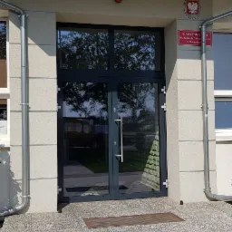 Szklane drzwi wejściowe do budynku z oknami po bokach, na zewnątrz rośliny w donicach i tabliczka informacyjna.