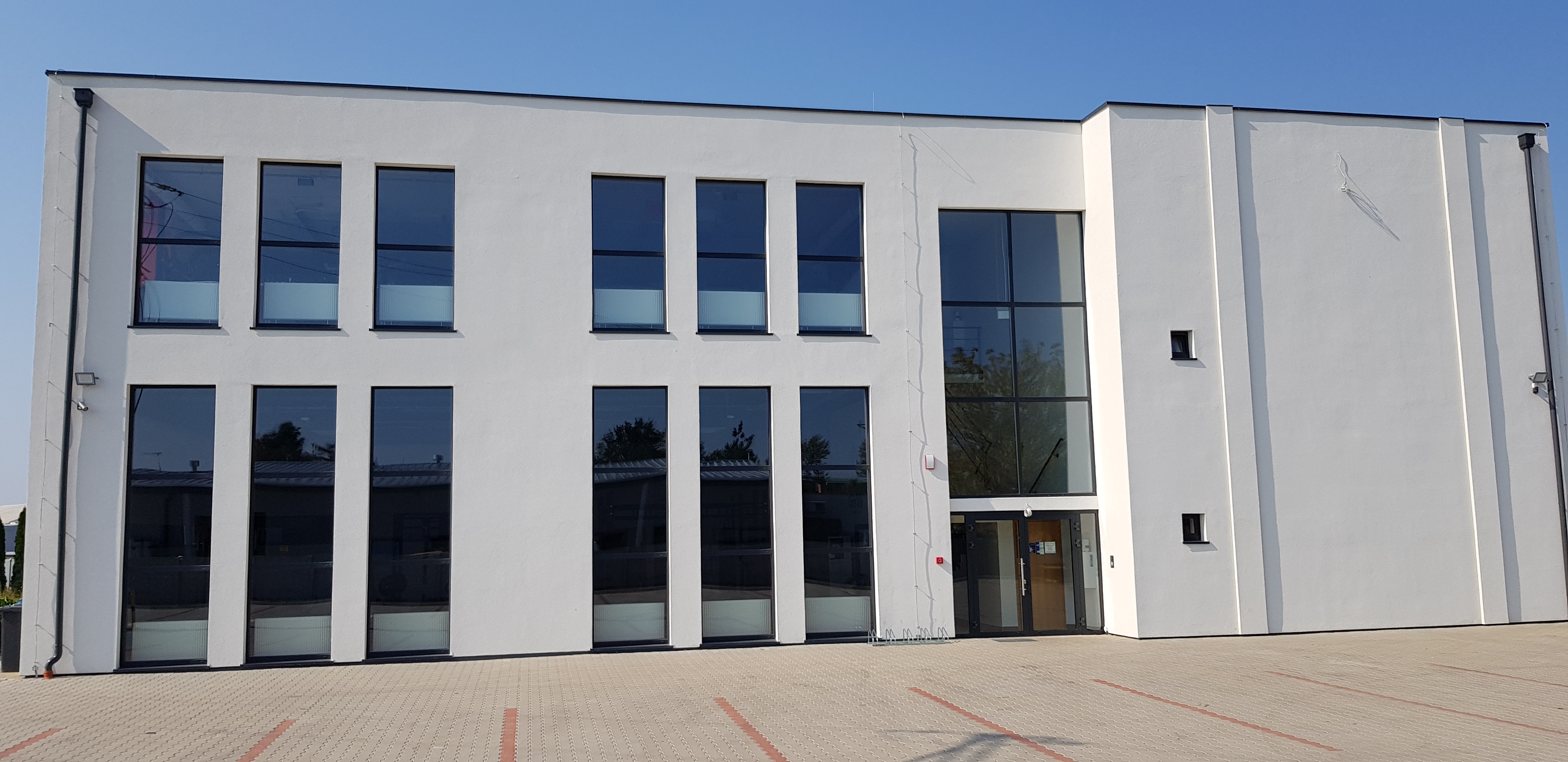 Nowoczesny budynek biurowy z dużymi oknami w czarnych ramach, widoczny chodnik z kostki brukowej i błękitne niebo.