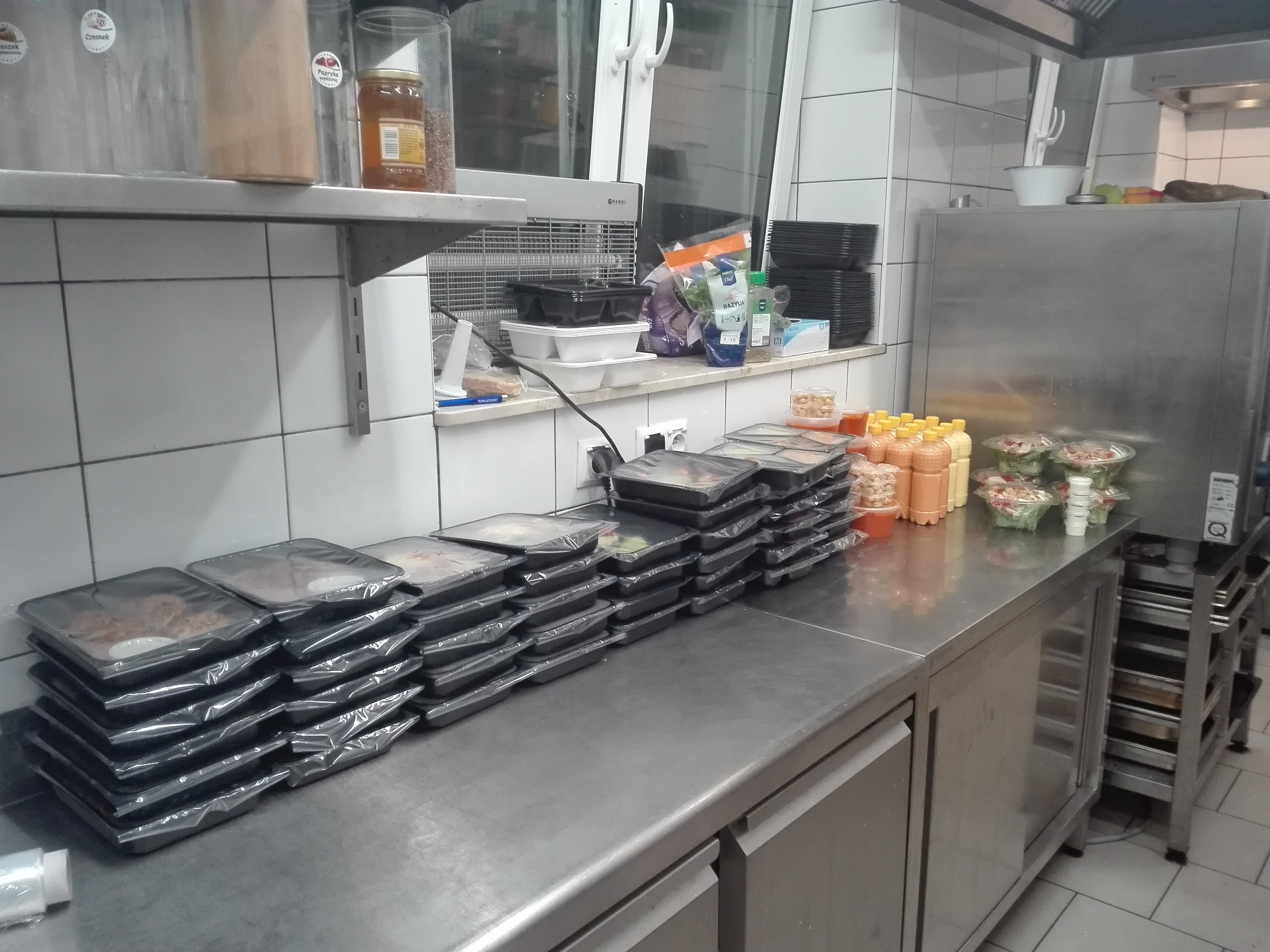 Przygotowane porcje dań w czarnych, szczelnie zamkniętych pojemnikach, ułożone w rzędach na metalowym blacie w kuchni, obok butelki z sosem i sałatki w przezroczystych pojemnikach.