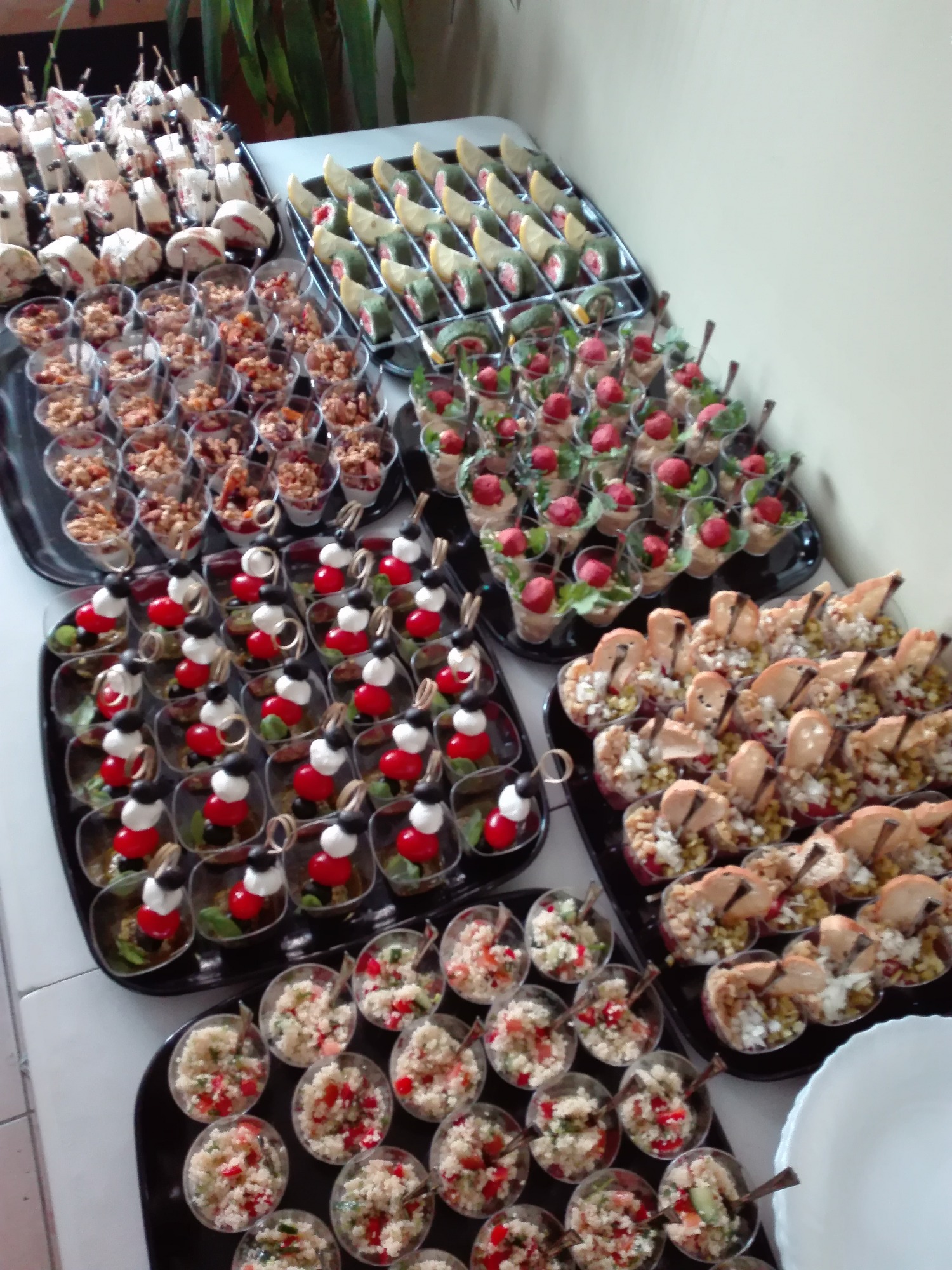 Różnorodne przekąski w małych naczyniach, ułożone na czarnych tacach: sałatki z kuskusem, koreczki z mozzarelli i pomidorów, roladki z łososiem i pastą, musli z owocami, przystawki z grzybami...