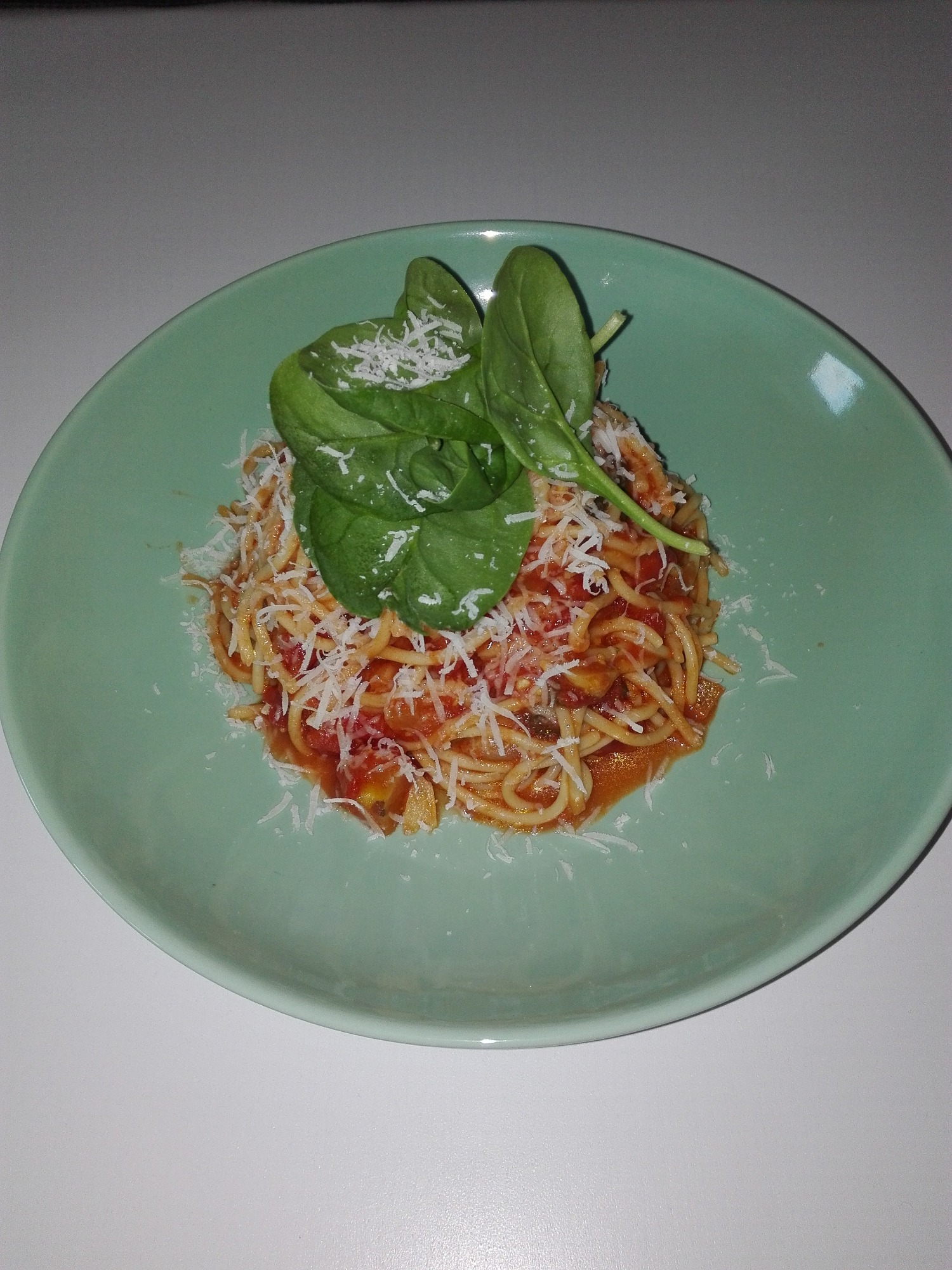 Porcja spaghetti z sosem pomidorowym, posypana parmezanem i udekorowana świeżymi liśćmi szpinaku, podana na turkusowym talerzu.