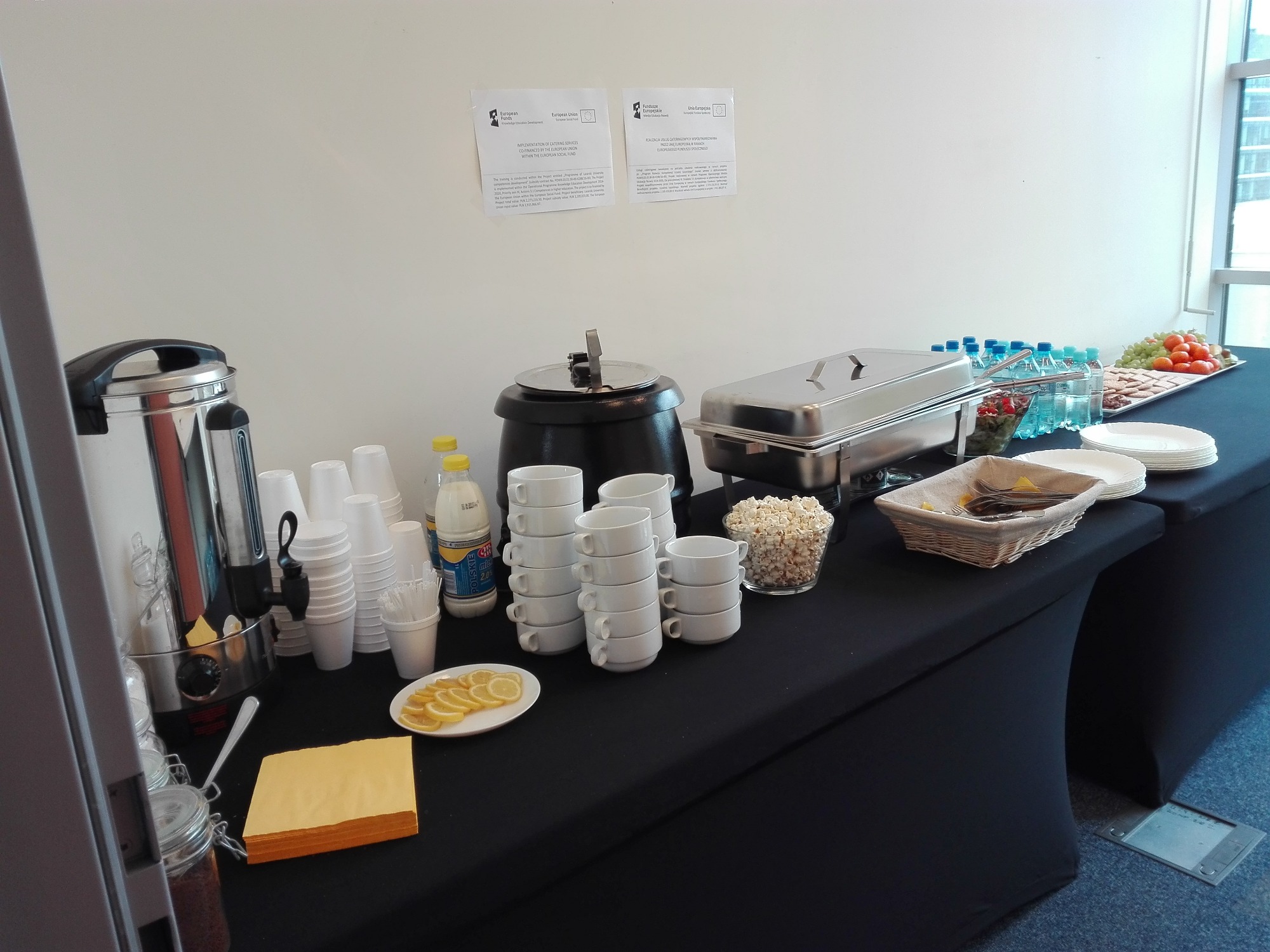 Stół cateringowy z czarnym obrusem, na którym znajdują się: termos do kawy, stosy białych kubków i filiżanek, butelka mleka, plasterki cytryny, naczynie z zupą, podgrzewacz do potraw, popcorn...