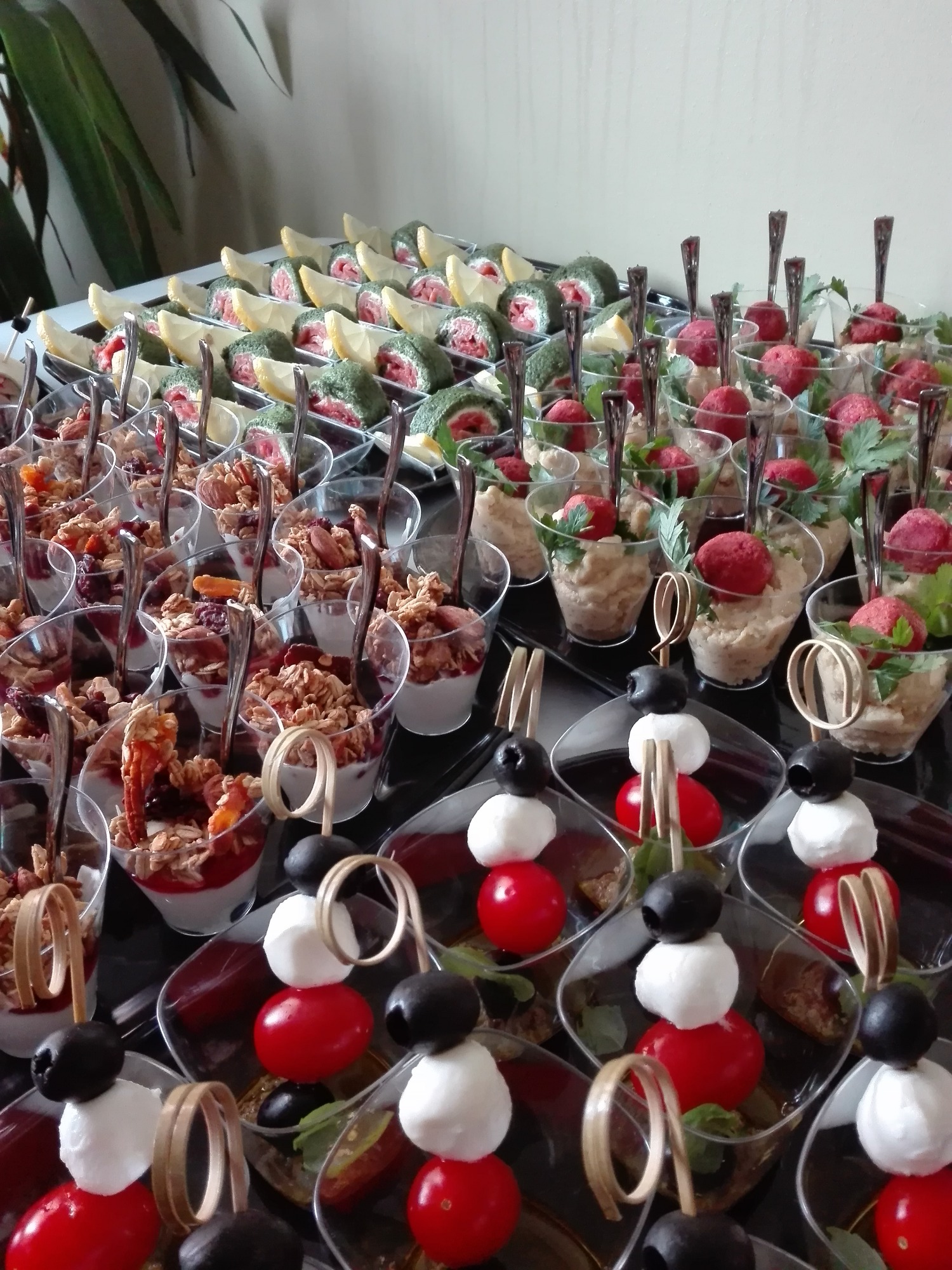 Elegancki catering: różnorodne przekąski w małych naczyniach, w tym szpinakowe roladki z łososiem i cytryną, mini-desery z granolą i jogurtem, kulki z mozzarelli, pomidorów i oliwek na wykałaczkach...