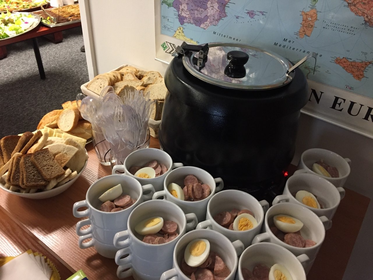 Bufet z zupą w czarnym garnku, kromkami chleba i porcjami zupy kiełbasianej z jajkiem w białych kubkach na tle mapy Europy.