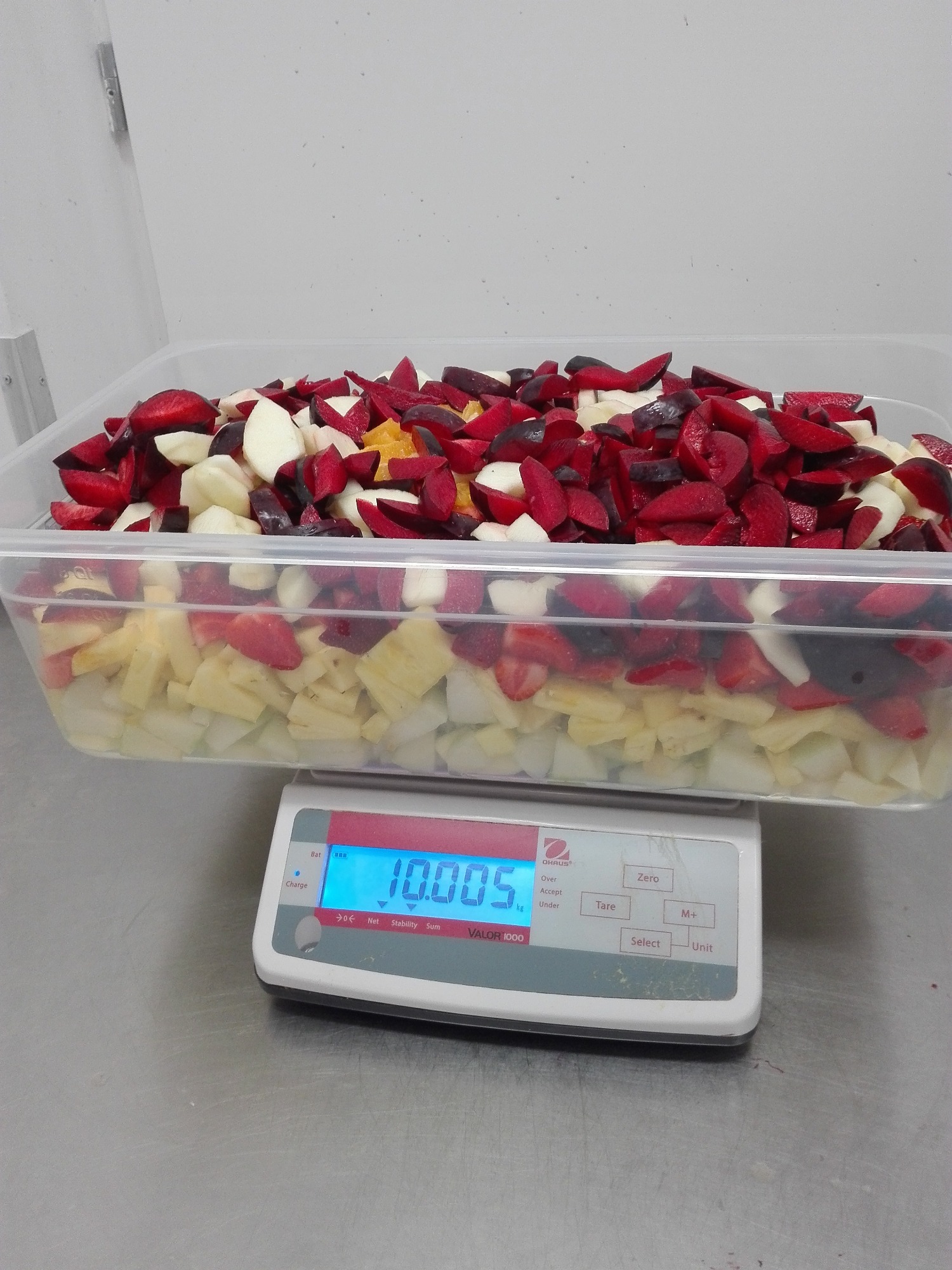 Pokrojone owoce, głównie śliwki, jabłka, ananasy i pomarańcze, w przezroczystym pojemniku na wadze elektronicznej wskazującej 10.005 kg, przygotowywane prawdopodobnie do sałatki owocowej.