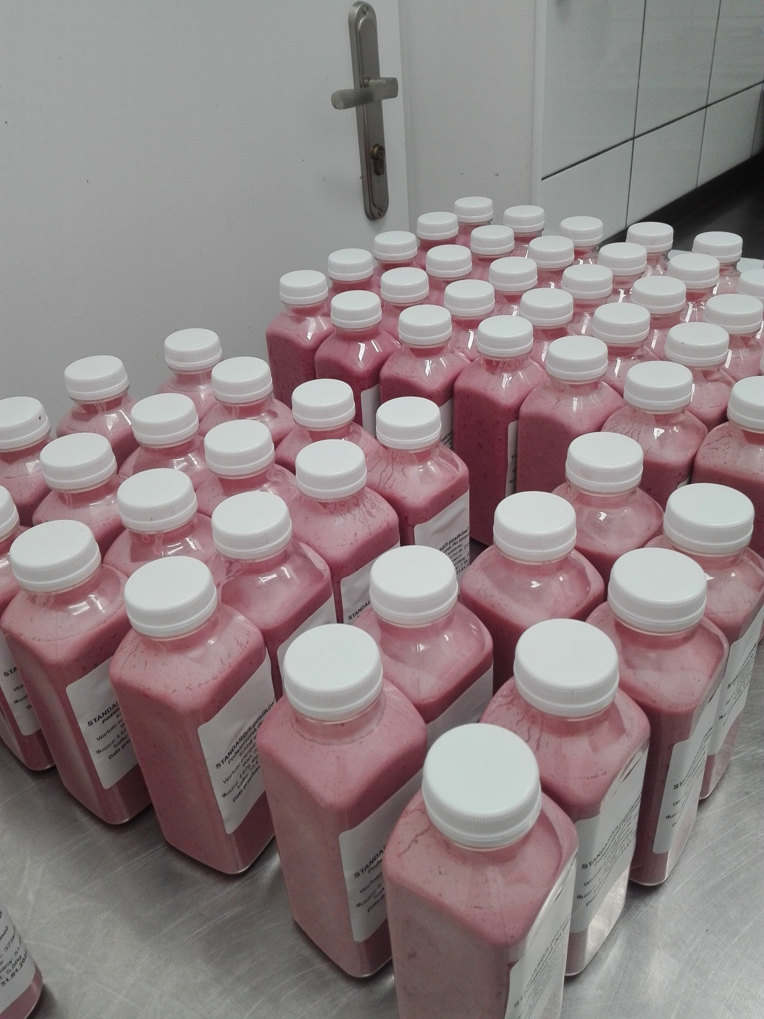 Rządki plastikowych butelek z różowym koktajlem, etykiety z informacjami o wartościach odżywczych, przygotowane do transportu na metalowym blacie w chłodnym pomieszczeniu.