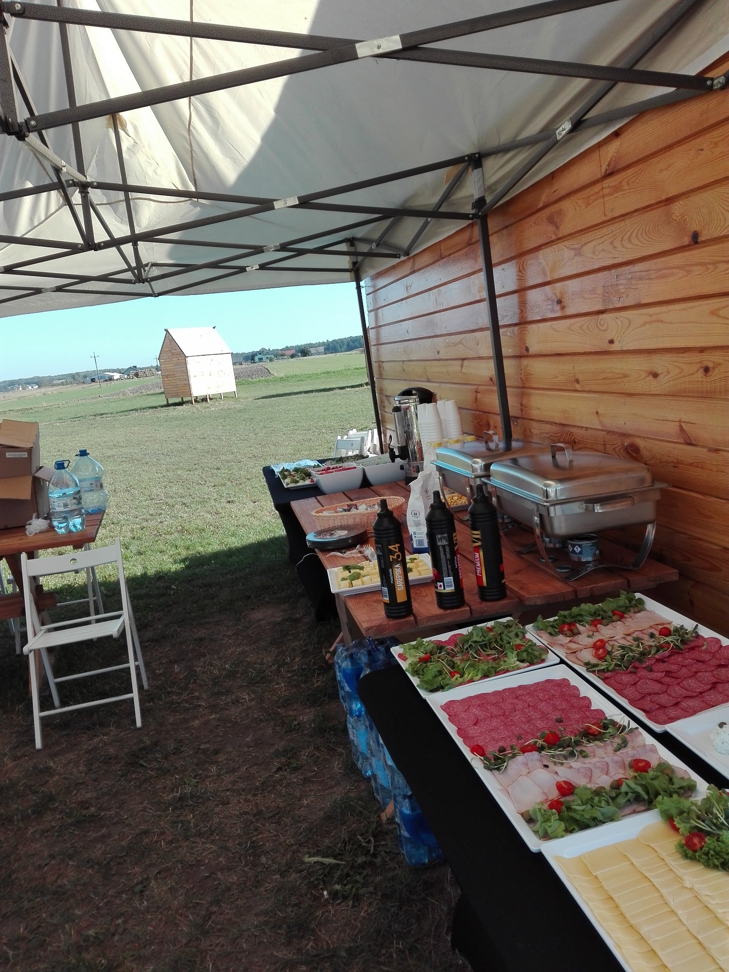 Zadaszony stół cateringowy na zewnątrz z półmiskami wędlin, serów i sałatek, butelkami sosów i podgrzewaczami do potraw; w tle pole i mała drewniana konstrukcja.