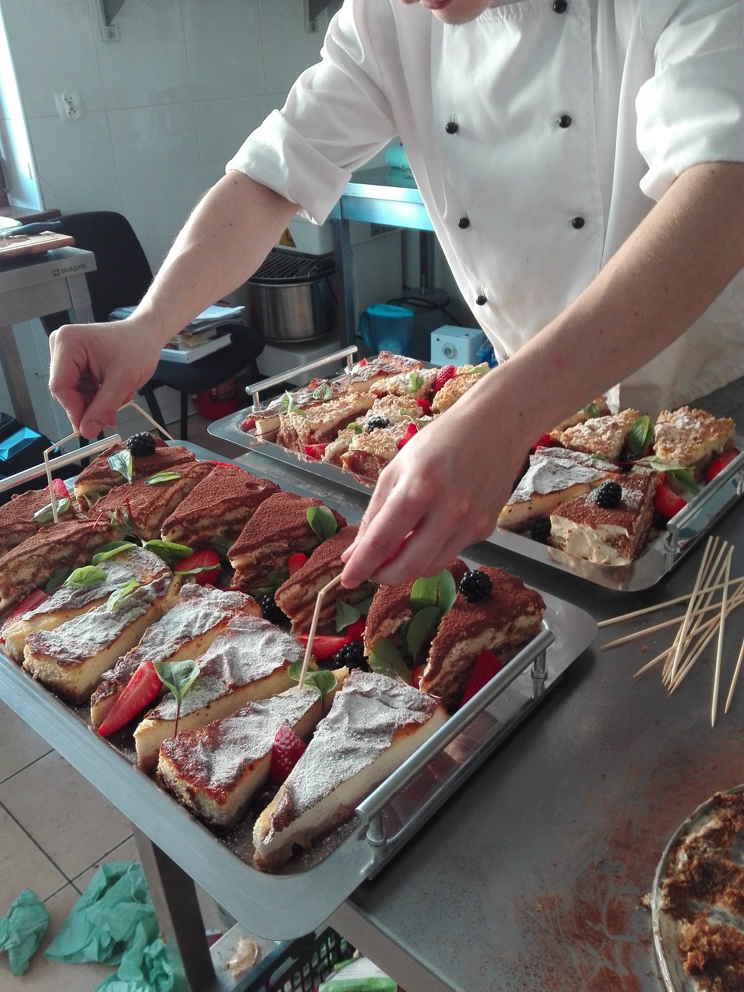 Kucharz w białym uniformie dekoruje srebrną tacę z porcjami ciast, w tym sernika posypanego cukrem pudrem i tiramisu z truskawkami i jeżynami, przygotowując je do serwowania.