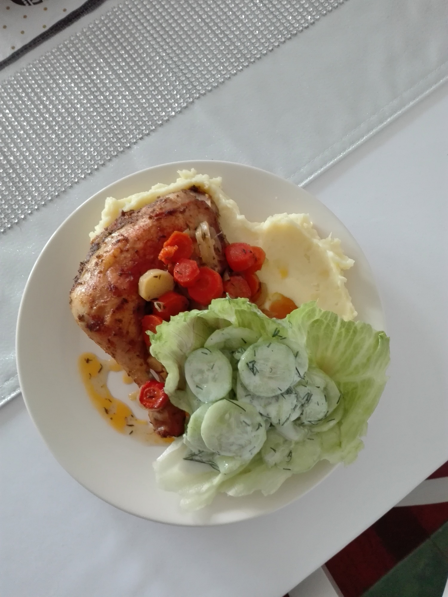 Upieczona noga kurczaka z dodatkiem puree ziemniaczanego, podsmażonych pomidorków cherry i sałatki z ogórków na liściach sałaty, podana na białym talerzu z dekoracyjną, połyskującą podkładką.