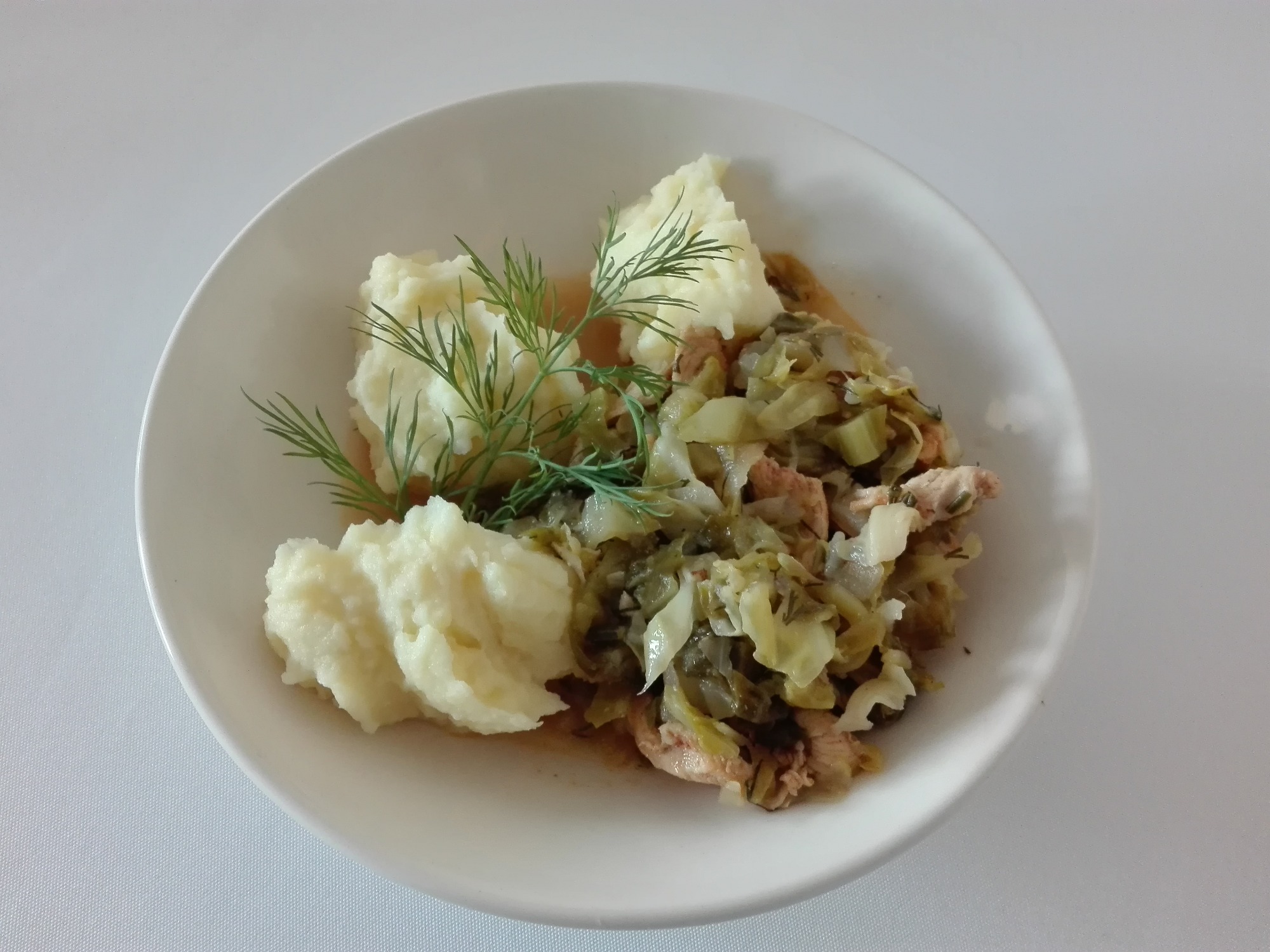 Porcja gotowanego mięsa z kapustą i ziemniaczanym puree, udekorowana gałązką świeżego koperku, podana na białym talerzu.