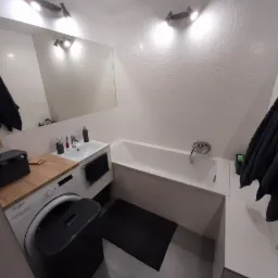 Jasna łazienka po remoncie z białą wanną, umywalką z szafką, pralką z drewnianym blatem i ręcznikami w kolorze grafitowym, ściany wyłożone białymi płytkami z delikatnym wzorem roślinnym.