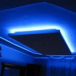 Sufit podwieszany z oświetleniem LED w kolorze niebieskim, centralny panel z czterema punktami świetlnymi, dekoracyjne listwy LED wzdłuż krawędzi sufitu, widoczny fragment pomieszczenia z zasłonami...