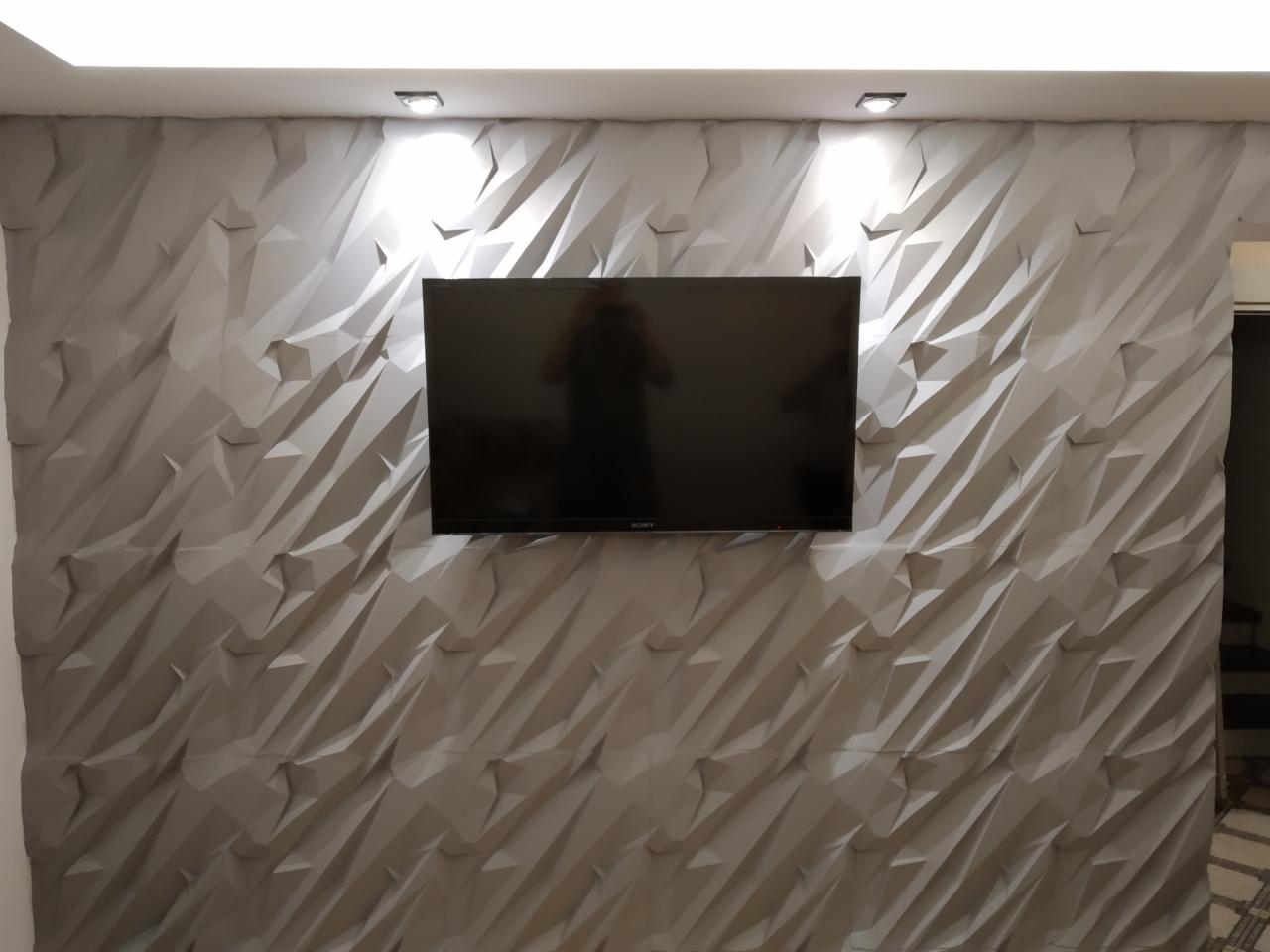 Szara ściana z trójwymiarową, geometryczną fakturą, na której wisi czarny telewizor Sony, oświetlona dwoma punktowymi lampami sufitowymi.