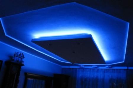 Sufit podwieszany z oświetleniem LED w kolorze niebieskim, centralny panel z czterema punktami świetlnymi, dekoracyjne listwy LED wzdłuż krawędzi sufitu, widoczny fragment pomieszczenia z zasłonami...