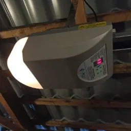 Zamontowany napęd do bramy garażowej z widocznym panelem sterowania i oświetleniem LED, zamocowany do konstrukcji dachu z blachy falistej i drewnianych belek.