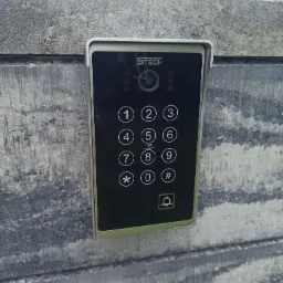 Panel domofonu z klawiaturą numeryczną i kamerą, zamontowany na szarej, betonowej ścianie o widocznej strukturze.