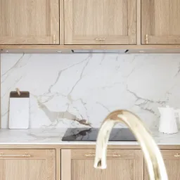 Jasna kuchnia z drewnianymi szafkami, blatem imitującym marmur i złotą baterią, widoczna płyta indukcyjna i czajnik.