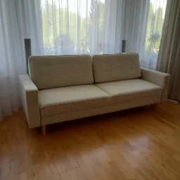 Jasna sofa z guzikami na oparciu, ustawiona na parkiecie przed oknem z białymi zasłonami, po renowacji.