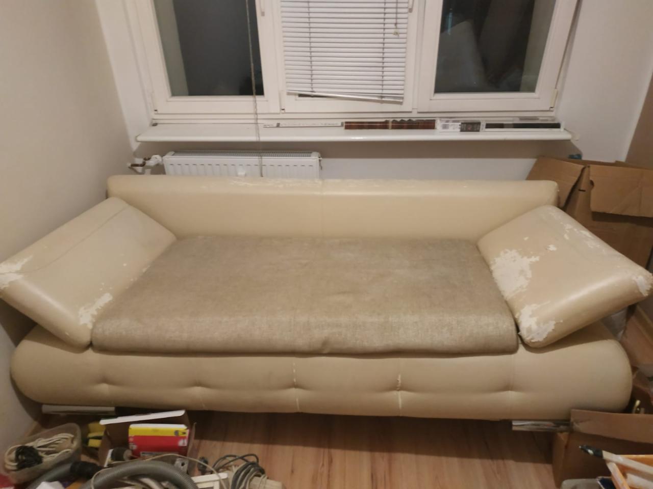 Beżowa sofa z widocznymi śladami użytkowania na podłokietnikach, jasny materiał siedziska, ustawiona pod oknem z żaluzjami, w tle kartony i przedmioty na podłodze.